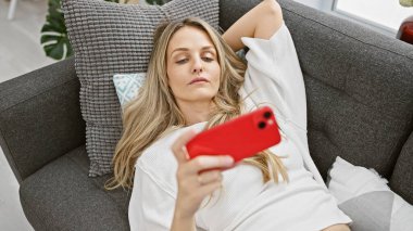 Sarışın bir kadın evdeki kanepede uzanıyor, kırmızı bir akıllı telefon kullanıyor, rahatlığı somutlaştırıyor ve modern bir ev hayatı yaşıyor..