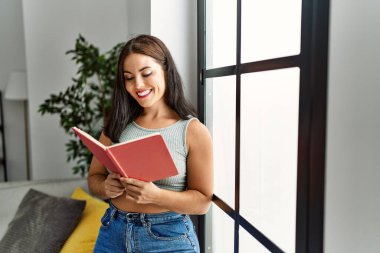 Young beautiful hispanic woman smiling confident reading book at home