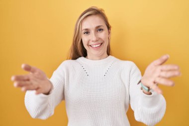 Beyaz süveter giyen, sarı arka planda gülümseyen, yardım eli uzatan beyaz bir kadın.. 