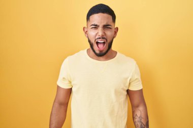 Young hispanic man standing over yellow background angry and mad screaming frustrated and furious, shouting with anger. rage and aggressive concept. 