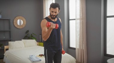 Sakallı, orta yaşlı yakışıklı bir adam modern bir yatak odasında dambıllarla çalışıyor..