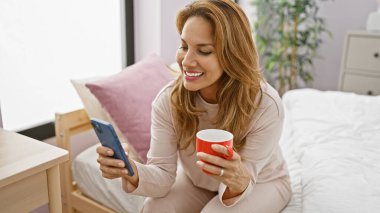 Gülümseyen İspanyol kadın, rahat bir yatak odasında akıllı telefonuna bakarken sabah kahvesinin tadını çıkarıyor.