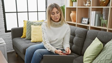 Güzel dekore edilmiş bir oturma odasında modern bir kanepede otururken dizüstü bilgisayar kullanan olgun sarışın bir kadın.