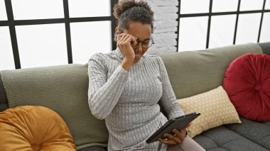 Genç Afrikalı Amerikalı bir kadın gözlüğünü ayarlıyor, tablet okuyor, kanepede rahatça oturuyor..