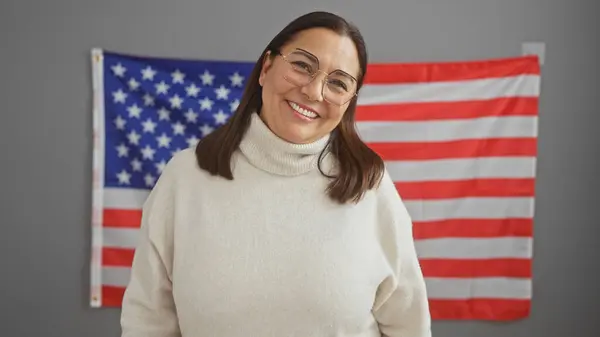 穿着白色毛衣的中年惊慌失措的女人站在办公室的一面美国国旗前微笑 — 图库照片