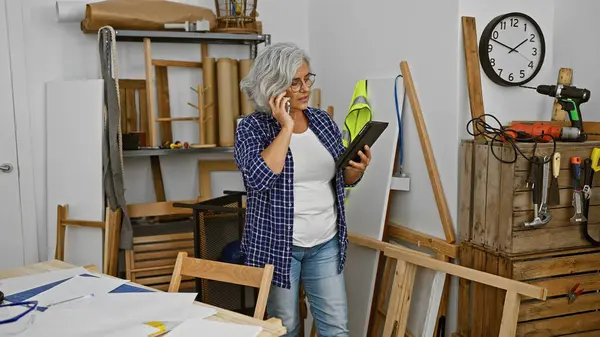 stock image Mature woman using tablet and phone in a well-equipped carpentry workshop.