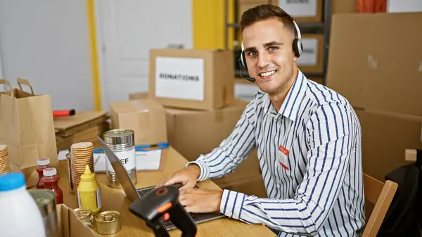 一个穿着条纹衬衫 头戴耳机的英俊年轻人在一个装有盒子的仓库里的计算机上工作 — 图库照片