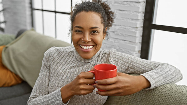 Улыбающаяся женщина, держащая красную кружку, сидящая на диване в уютной гостиной, излучающая расслабление и тепло.