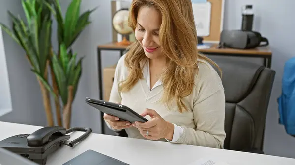 stock image A young hispanic woman engages with a tablet in a modern office setting, exuding professionalism and focus.