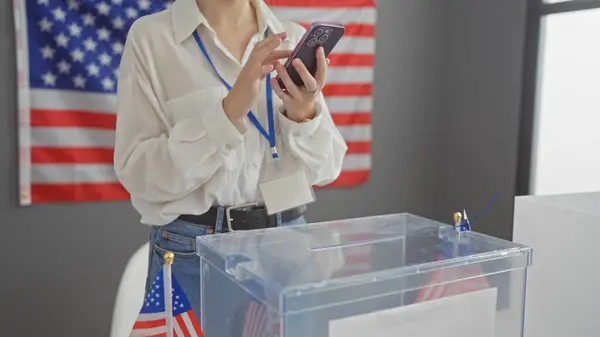 ラニヤードを持つ若い女性は 投票センターで彼女の携帯電話をチェックし 私たちの旗と投票箱 — ストック写真