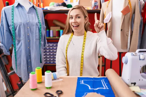 Young caucasian woman dressmaker designer working at atelier smiling with an idea or question pointing finger up with happy face, number one