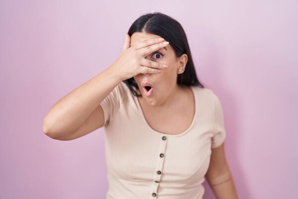 Молодая латиноамериканка, стоящая на розовом фоне, заглядывая рукой в лицо и глаза, смотрит сквозь пальцы со смущенным выражением лица.. 
