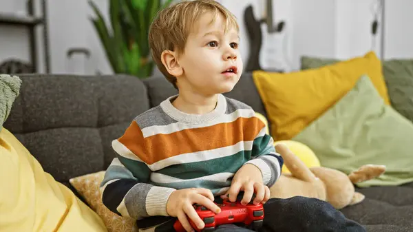 一个全神贯注的金发男孩在一个舒适的客厅里和一个游戏控制器玩耍 四周都是五颜六色的枕头和一个毛绒玩具 — 图库照片