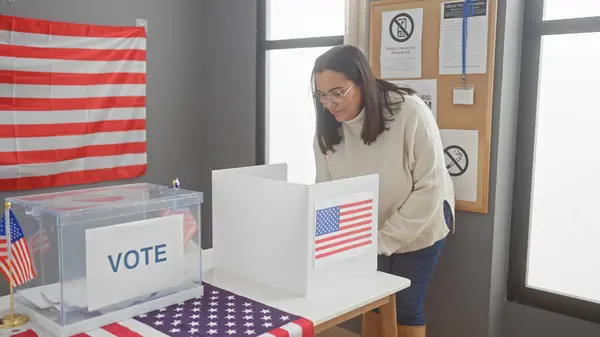 stock image A middle-aged hispanic woman votes indoors in a usa electoral center with an american flag visible.
