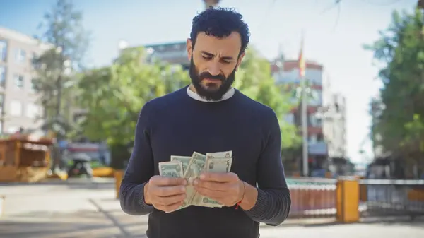 A mature, bearded man counts money outdoors on a sunny city street, portraying everyday financial transactions.