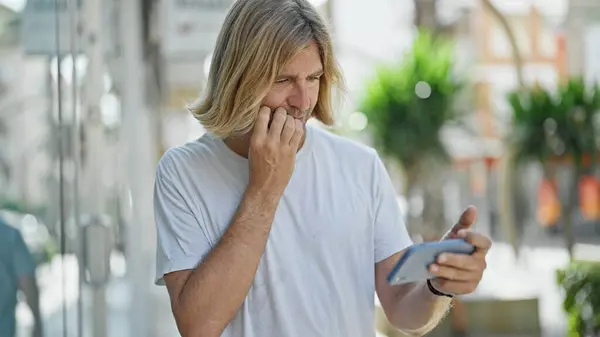 一个头发长长的困惑男人在阳光灿烂的城市街道上拿着电话 — 图库照片