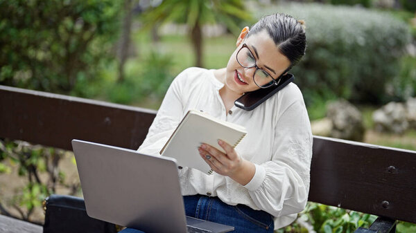 Молодая красивая латиноамериканка, разговаривающая по телефону, пишет на блокноте, сидя на скамейке в парке