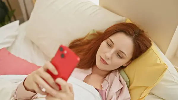 一位年轻的高加索女人一边在卧室里放松下来 一边在智能手机上浏览 这让人产生了休闲和连接的感觉 — 图库照片