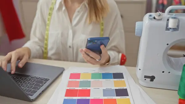 A young woman in a tailoring atelier selects fabric colors while holding a smartphone, with a sewing machine and laptop present.