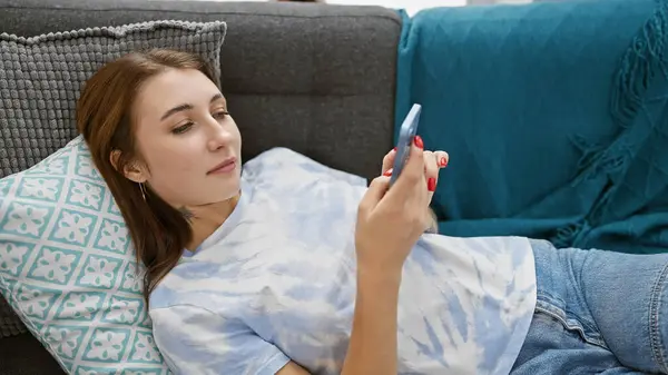 若いブルネットの白人女性は 居心地の良い家の設定で彼女のスマートフォンを使用して 屋内ソファーでリラックス — ストック写真