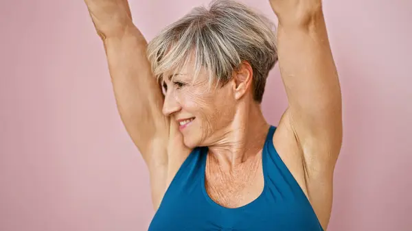A smiling mature woman with short grey hair in a blue tank top stretching her arms against a pink background.