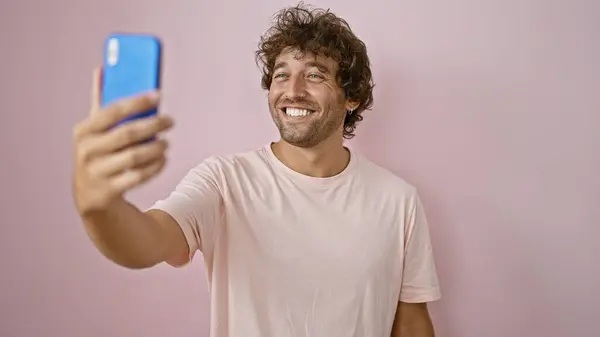一个快乐的年轻人拿着一个蓝色的智能手机 靠着一堵粉红色的墙 带着一个自拍 散发着自信和欢乐 — 图库照片