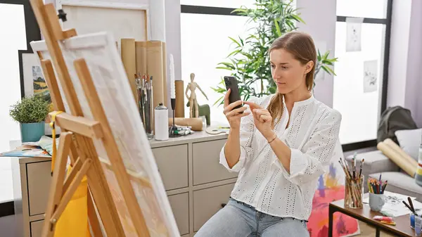 A focused caucasian woman artist in a studio uses a smartphone beside her easel and art supplies.