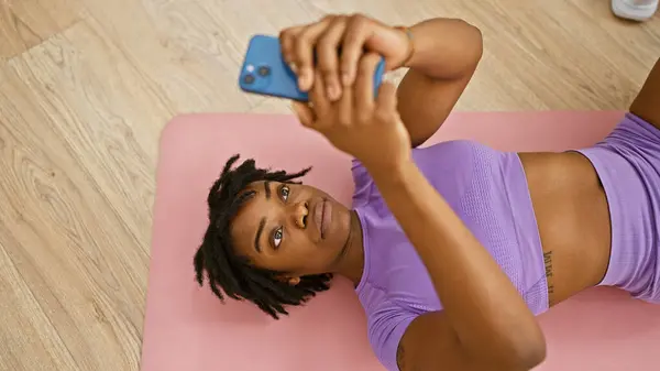 African american woman lying on a yoga mat indoors taking a selfie with her smartphone.