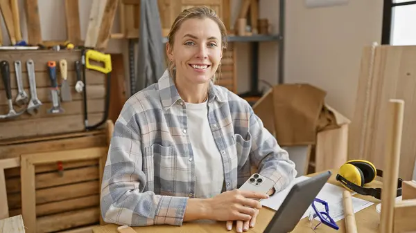 一个穿着格子呢衬衫 面带微笑的高加索女人坐在一个木匠车间里 手里拿着工具和木头 — 图库照片