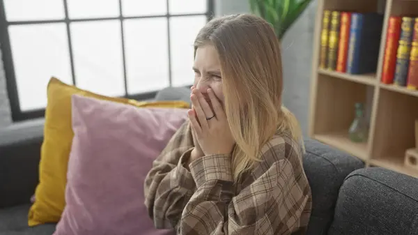 stock image A young blonde woman appears distressed sitting indoors, her hands covering her mouth, with plush pillows and a bookshelf in the background.