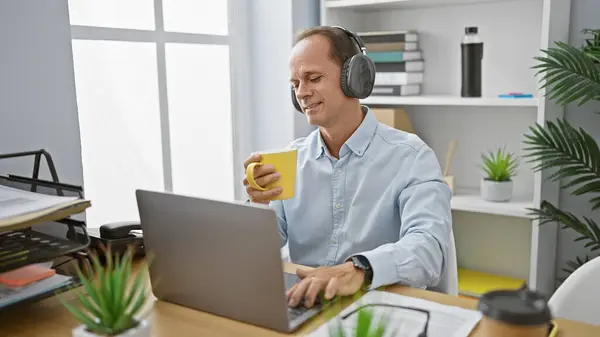 Cheerful middle age man, a successful executive, joyfully working at his office desk, listening to music and sipping his morning espresso coffee, brightening up the indoor workplace.