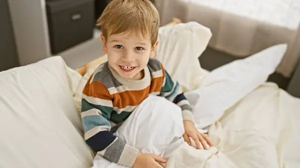 stock image A cheerful blond toddler boy sitting in a white bed within a cozy room, looking directly at the camera.