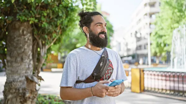 Smiling hispanic man with beard and bun hairstyle texting on smartphone in park