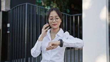 Gözlüklü Asyalı kadın akıllı telefon kullanıyor ve şehirde saatleri kontrol ediyor..