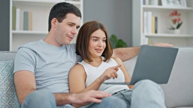 Güzel bir çift kanepede oturup dizüstü bilgisayar kullanarak evde gülümsüyor.