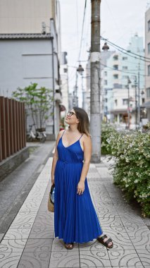 Gözlüklü güzel İspanyol kadın, sıradan bir şekilde ayakta ve mutlu bir şekilde gülümsüyor, Tokyo 'nun modern sokaklarında kendine güvenini sergiliyor, etrafına bakıyor, Japonya' nın kentsel manzarasının tadını çıkarıyor..