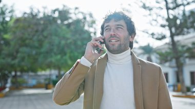 İspanyol adam telefonda konuşuyor, açık gökyüzünün altındaki yeşil park manzarasının ortasında ışıldıyor..