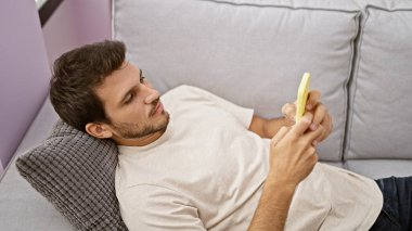 Rahat bir genç adam, rahat bir oturma odasındaki kanepede uzanırken akıllı telefonuyla mesaj atıyor..