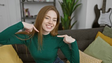 Keyifli genç bir kadın arka planda gitarıyla rahat bir oturma odasında kendini işaret ediyor.