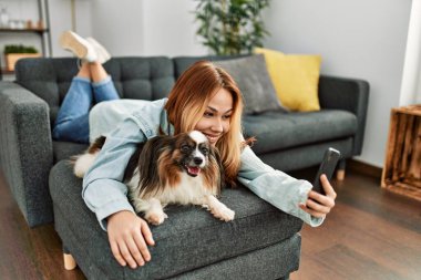 Beyaz bir kadın köpeğe sarılıyor ve evde akıllı telefondan selfie çekiyor.