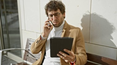 Gözlüklü şık bir adam şehirde otururken telefon ve tabletle birden fazla iş yapıyor..