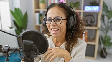 Radyo stüdyosunda konuşan, profesyonel yayın yapan bir atmosferi canlandıran, kulaklıklı gülümseyen bir kadın..