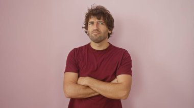 A skeptical young hispanic man with a beard is standing arms crossed against a pink wall, conveying a sense of contemplation. clipart