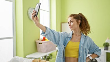 Kendine güvenen genç bir kadın, parlak bir çamaşır odasında akıllı telefonuyla selfie çekiyor..