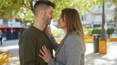 Kadın, güneşli bir sokakta yan yana duran bir erkeğin yüzüne sevgiyle dokunur..