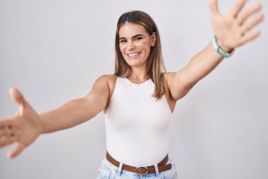 Latin kökenli genç bir kadın beyaz arka planda durup kameraya bakarak sarılmak için kollarını açmış gülümsüyor. Mutluluğu kucaklayan neşeli bir ifade.. 