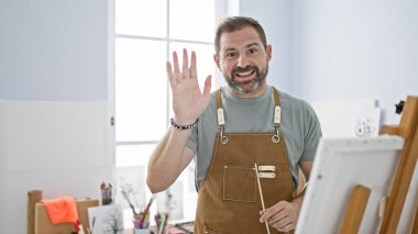 Beyaz saçlı, önlüklü İspanyol olgun bir adam. Parlak bir sanat stüdyosunda el sallıyor..