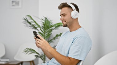 Genç sakallı bir adam akıllı telefonunu kullanarak modern bir bekleme odasında kulaklıkla müzik dinliyor..