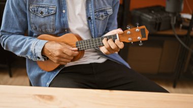 Hispanic man playing ukulele indoors, wearing denim, seated by a wooden table in a casual studio setting. clipart