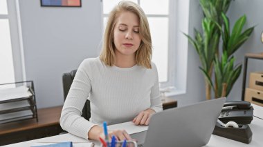 Sarışın, kısa saçlı bir iş kadını modern ofis ortamında bilgisayarının başında çalışmaya odaklanmış..